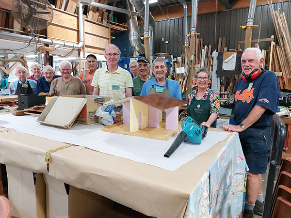 group standing in front of table in a shed. Items on the table include a minature house and a leaf blower