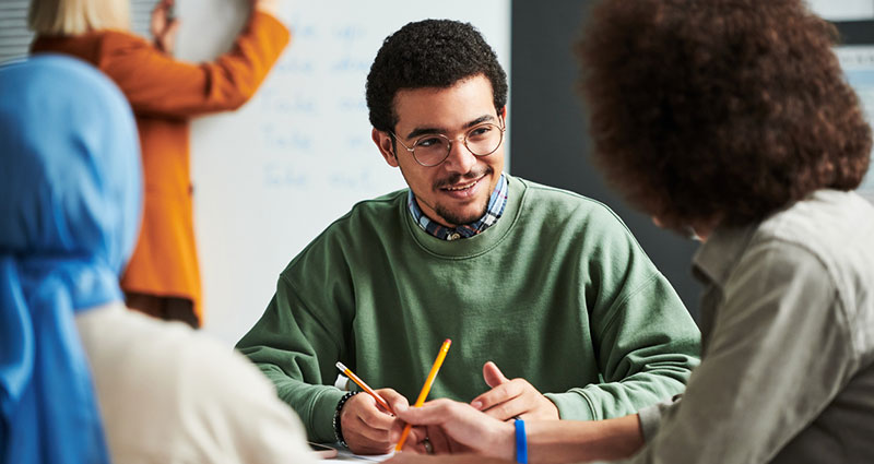 Happy young man in eyeglasses communicating with another student at break between lessons against teacher preparing for presentation