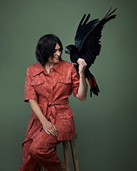 Leila in red coloured outfit sitting on chair with black cockatoo on her shoulder