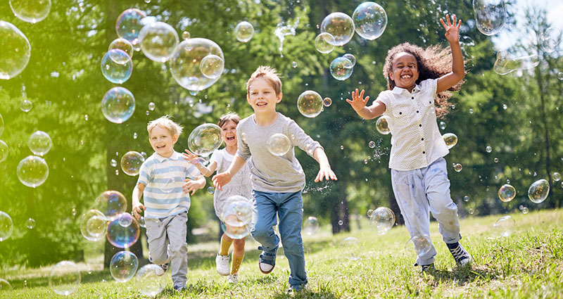 Multi-ethnic group of little friends with toothy smiles on their faces enjoying warm sunny day while participating in soap bubbles show