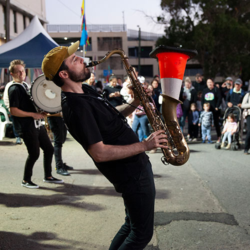 man playing saxaphone (with upside down traffic cone inserted) in front of crowd